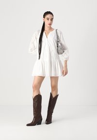 Rochie albă, brodată, cu mâneci lungi, cu fustă în straturi și decolteu în V. Botine cowboy maro, până la genunchi, cu detalii complicate. Geantă de mână argintie.