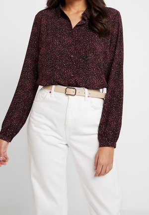 Femme portant un chemisier à manches longues à motif rouge foncé, rentré dans un pantalon blanc taille haute avec une ceinture beige clair avec boucle.