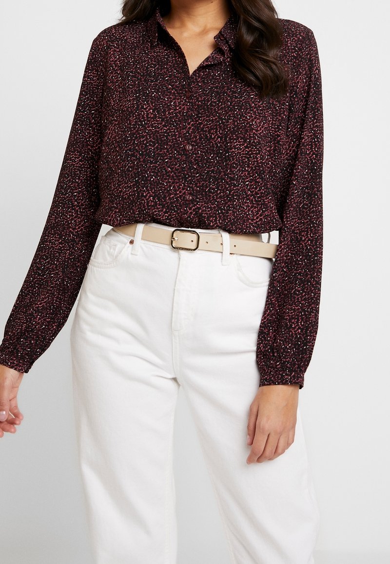 Femme portant un chemisier à manches longues à motif rouge foncé, rentré dans un pantalon blanc taille haute avec une ceinture beige clair avec boucle.