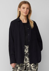 Zwarte textuurshirt met een knoopsluiting aan de voorkant, lange mouwen en een borstzak, gecombineerd met gestreepte crèmekleurige en zwarte broek.