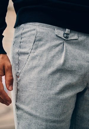 Grau gemusterte Hose mit sichtbarem Knopfverschluss, Seitentasche und strukturiertem Stoff. Figurbetonte Passform mit dezentem Faltenwurf an der Taille.