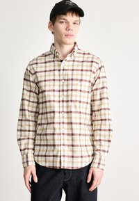 Camicia a quadri beige con striscie color bordeaux, gialle e bianche, colletto button-down, maniche lunghe e taschino sul petto. Indossata con jeans scuri.