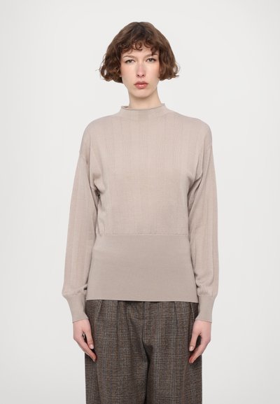 Beige ribbeltrui met een hoge halslijn en lange mouwen, met een strak lager gedeelte en een zachte textuur. Gepaard met bruine geruite broek.
