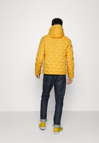 Gul vadderad jacka med huva, med en texturerad yta och en figursydd design, tillsammans med mörka jeans och gula sneakers.