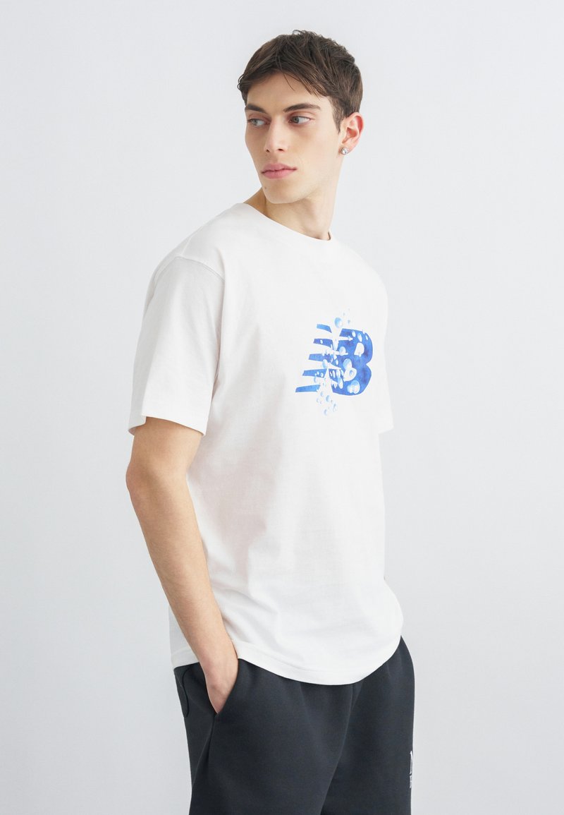 Jeune homme en t-shirt blanc avec un logo "NB" bleu et pantalon noir, debout les mains dans les poches, regardant vers la gauche sur un fond uni.