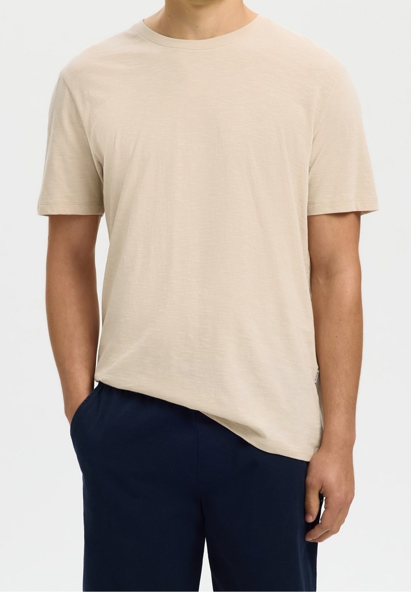 Homme portant un t-shirt beige uni à manches courtes avec un pantalon bleu marine, une main dans la poche, sur un fond clair uni.