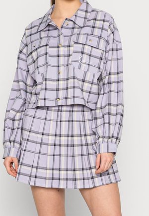 Femme portant une veste courte à carreaux lavande avec des poches poitrine boutonnées, assortie à une mini-jupe plissée.