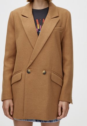 Woman wearing a camel double-breasted wool blazer with notch lapels, two front pockets, and button details over a graphic T-shirt.