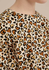 Bavlněná košile s béžovým pozadím a černým a hnědým leopardím vzorem ve tvaru srdce. Design s kulatým výstřihem, měkká textura.