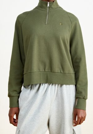 Persoon draagt een olijfgroene half-rits trui met geribbelde manchetten en zoom, gecombineerd met lichtgrijze losse broek.