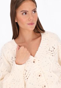 Tânără cu păr lung, castaniu, îmbrăcată într-un cardigan tricotat de culoare crem, cu pete multicolore și nasturi, privind spre dreapta.