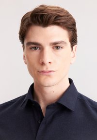 Młody mężczyzna z krótkimi brązowymi włosami, ubrany w ciemnogranatową koszulę zapinaną na guziki, stoi frontalnie z neutralnym wyrazem twarzy na jasnym tle.