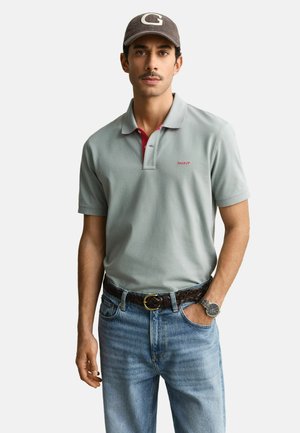 Man draagt een grijs poloshirt, blauwe spijkerbroek, bruine gevlochten riem, grijze pet met letter G en polshorloge, staat met één hand in de zak.