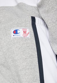 Sudadera gris con un panel blanco, acentos en azul marino y una etiqueta del logo de Champion con un borde rojo y detalles en texto. Fabricada con un tejido suave.