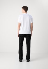 T-shirt bianca in cotone con maniche corte, colletto a giro, indossata con pantaloni neri dritti e scarpe nere, vista di spalle.