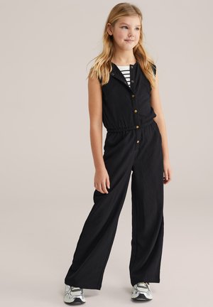 Salopetă neagră fără mâneci, cu finisaj texturat, nasturi în față și talie elastică. Purtați peste un tricou negru cu dungi albe, asortată cu adidași.