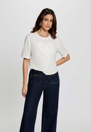 Frau trägt ein weißes kurzärmeliges Shirt mit strukturiertem Herzdesign und dunkle Jeans mit weitem Bein, steht mit der Hand in der Hüfte vor einfarbigem Hintergrund.