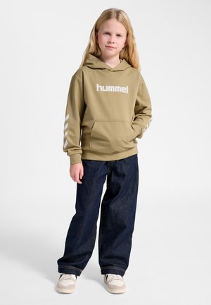 Ung pige med langt blondt hår iført en beige hummel-hoodie, mørke vide jeans og hvide sneakers, stående mod en ensfarvet baggrund.