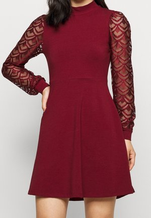 Frau in einem burgunderroten Kleid mit einem einfarbigen Oberteil und durchscheinenden langen Ärmeln aus Spitze mit geometrischen Mustern.