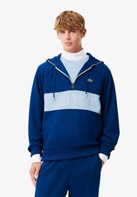 Marineblå hettegenser med lyseblå horisontal stripe som har "LACOSTE" logo, frontlås og hvite ribbestrikkede mansjetter.