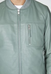 Světle zelená kožená bomber bunda s žebrovaným límcem, předním zipovým zapínáním a dvěma zipovými kapsami na hrudi a po stranách. Hladký povrch.