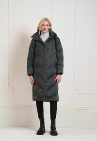 Manteau long matelassé de couleur gris foncé avec surpiqûres en forme de chevrons, col montant et devant boutonné. Porté par-dessus un col roulé gris clair et un pantalon noir.