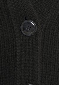 Tmavý pletený kardigan s texturovaným povrchem a velkým kulatým černým plastovým knoflíkem se čtyřmi dírkami.