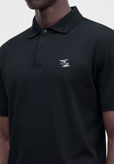 Homme portant un polo noir à manches courtes avec un col boutonné et des initiales blanches brodées sur la poitrine gauche.