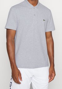Szara bawełniana koszulka polo z klasycznym kołnierzykiem, dwoma guzikami, krótkimi rękawami oraz zielonym logo Lacoste na lewej piersi.