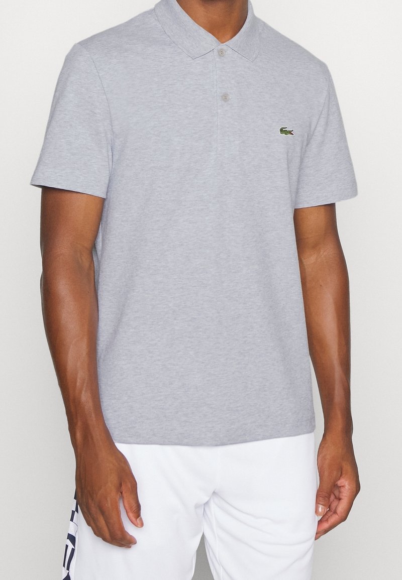 Szara bawełniana koszulka polo z klasycznym kołnierzykiem, dwoma guzikami, krótkimi rękawami oraz zielonym logo Lacoste na lewej piersi.