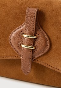 Brūna zamšāda rokassoma ar teksturētu ādas vākumu, zelta toņa metāla detaļām, dekoratīvu cilpu un šuvju apdari aizdarei.