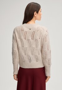 Strickpullover in hellem Beige mit offenen Mustern, geripptem Kragen und Bündchen, mit einem kleinen Logo-Detail auf der Rückseite. Ärmel in mittellanger Länge.
