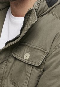 Detailní záběr osoby v olivově zelené bundě s knoflíkovou kapsou a bílém tričku pod ní.