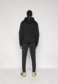 Μαύρο φούτερ με υφασματικό σχέδιο στα μανίκια, σε συνδυασμό με σκούρο παντελόνι με σχέδια και μπεζ ξύλινα παπούτσια. Απλός, λιτός σχεδιασμός συνολικά.