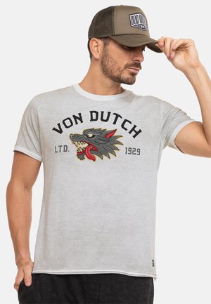Homme portant une casquette beige Von Dutch et un t-shirt gris clair Von Dutch avec un logo de loup grognant et le texte "LTD. 1929" à l'avant.