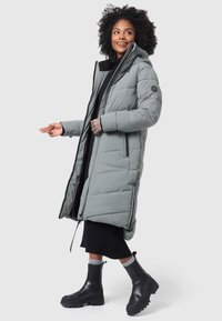 Manteau d'hiver long gris en duvet avec fermeture éclair, capuche et poignets côtelés. Porté par-dessus une jupe noire côtelée et assorti avec des bottines noires à semelle épaisse.