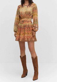 Vestido con estampado, de mangas largas, con cintura ceñida y falda acampanada en tonos naranjas, verdes y dorados, combinado con botas de ante marrones hasta la rodilla.