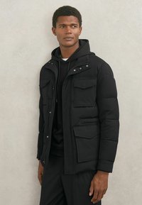 Man draagt een zwarte gewatteerde jas met capuchon en klepzakken, staande tegen een lichtgrijze muur, licht opzij kijkend.