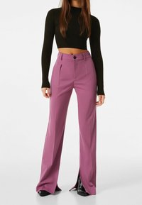 Kvinna i högmidjade mauve vida byxor med framtunga slitsar, tillsammans med en svart kort ribbad långärmad topp och svarta stövlar.