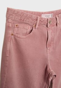 Svijetlo ružičaste traperice s glatkom teksturom, dizajnom s pet džepova, vidljivim šavovima i zlatnim dodacima na džepovima.