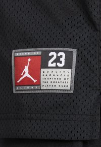 Czarna siateczkowa tkanina z metką z czerwonym i szarym logo, "23," oraz napisem "PRODUKTY JAKOŚCIOWE INSPIROWANE NAJWIĘKSZYM GRACZEM WSZYSTKICH CZASÓW."