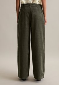 Persoon in een groene, hoge taille wijde broek met achterzakdetails, gecombineerd met een crèmekleurige gehaakte top, staand voor een neutrale achtergrond.