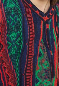 Värikäs neulottu neule, jossa on punaisen, vihreän ja sinisen sekoitus kuvioita, kohokuvioituja raitoja ja V-kaula-aukko.