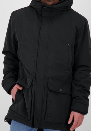 Man wearing black hooded winter jacket with flap pockets and snap buttons, hands partially in pockets, against plain background.