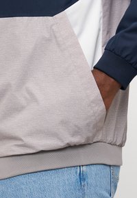 Chaqueta ligera gris con acentos en azul marino y paneles blancos. Cuenta con un tejido texturizado, puños elásticos y un bolsillo lateral. Denim visible abajo.