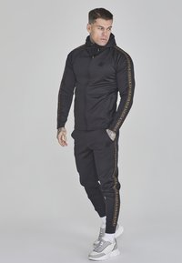 Schwarzer athletischer Trainingsanzug mit goldenem Logo entlang der Ärmel, taillierte Passform, Kapuzenjacke mit Reißverschluss und passenden Jogginghosen. Graue und weiße Turnschuhe.
