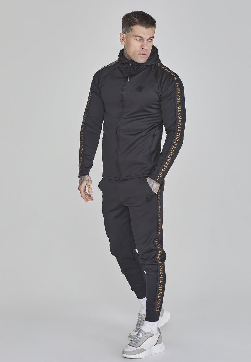 Schwarzer athletischer Trainingsanzug mit goldenem Logo entlang der Ärmel, taillierte Passform, Kapuzenjacke mit Reißverschluss und passenden Jogginghosen. Graue und weiße Turnschuhe.