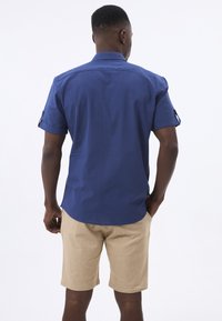 Camisa de manga corta de tejido azul oscuro con cuello y mangas enrolladas, combinada con pantalones cortos beige que tienen un acabado texturizado.