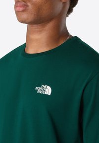 Nærbillede af person iført en mørkegrøn The North Face t-shirt med hvidt logo på brystet og en sølvfarvet kæde synlig om halsen.