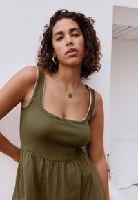 Olivengrøn ærmeløs kjole med ribbet tekstur, firkantet halsudskæring og hvid kant. Har en udvidet bund og er kombineret med guldsmykker.